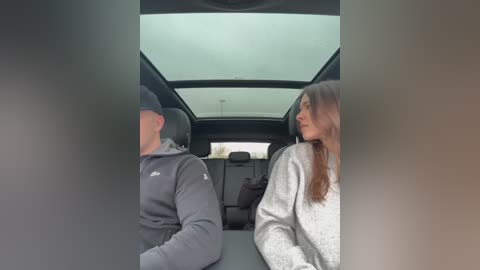 Video of a man and woman in a car. Man, wearing a gray hoodie, looks left; woman, with long hair, in a white sweater, gazes out the window.