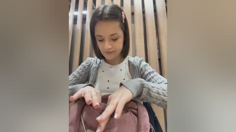 Video of a young girl with straight brown hair, wearing a white shirt, gray cardigan, and brown pants, seated on a wooden bench, focused on her hands, with a beige wall and wooden slats as background.