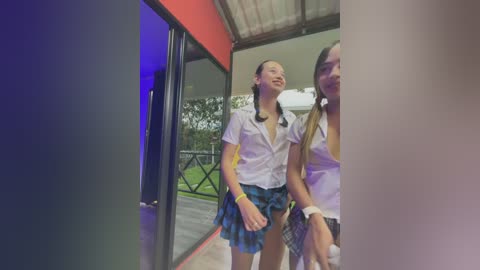 Video of two young women, one with long hair, in school uniforms, smiling, standing in a doorway, green grass background.