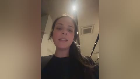 Video of a young woman with dark hair, smiling, wearing a black top, taken indoors with a dim, warm lighting. The background shows a plain room with a ceiling light and a vent.