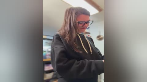 A video of a young woman with long, straight brown hair and glasses, wearing a black hoodie, standing in a dimly lit room with a TV in the background.