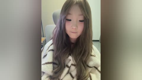 Video of a young Asian woman with long, wavy brown hair, wearing a beige striped robe, seated in an office chair with a grey backrest, blushing and looking down.