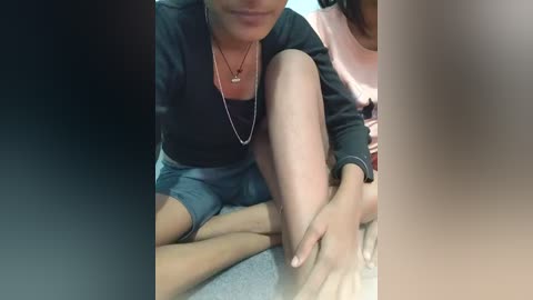 Media: Video of a woman with medium brown skin and a slender build, sitting cross-legged on a grey carpet. She wears a black top, silver necklace, and dark shorts. Her hands gently massage her own feet.
