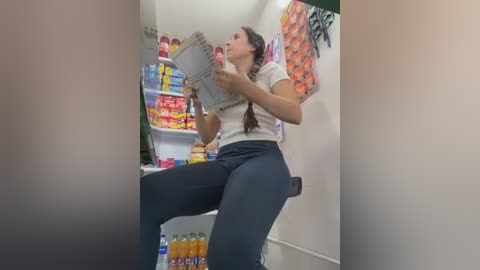 Video of a woman with long brown hair, wearing a white t-shirt and black pants, drinking from a bottle while standing on a chair in a cluttered kitchen with colorful snacks and beverages on shelves.
