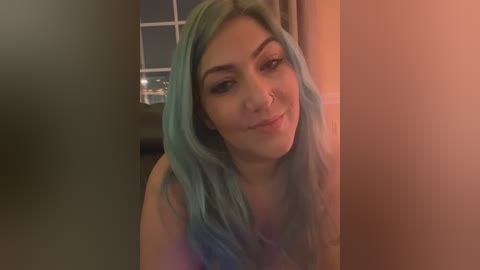 Video of a young woman with long, blue hair, fair skin, and a small nose piercing, smiling slightly. Background features a window with a view of a cityscape at night. Warm, dim lighting.