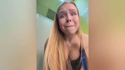A video of a young Caucasian woman with long blonde hair, wearing a blue tank top, in a green room, making a silly face.