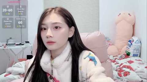 Video of an Asian woman with long black hair in a plush, pastel-colored bedroom, wearing a white jacket with colorful cartoon designs, sitting on a bed with a large stuffed animal.