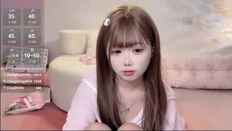 Video of a young Asian woman with long brown hair, wearing a pink cardigan, lying on a bed with a white blanket. Background shows a digital screen displaying weather and temperature information.