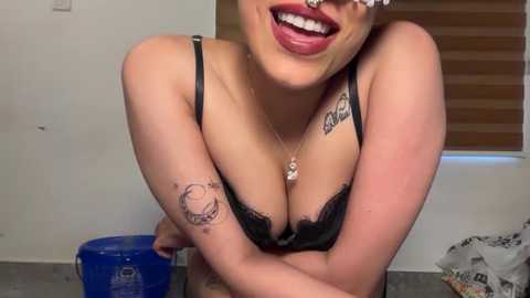 Video of a smiling, light-skinned woman with tattoos, wearing black lace bra, leaning against a blue bucket in a cluttered room with a brown window blind.