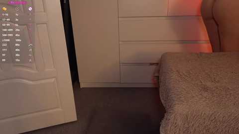 Video of a bedroom with beige carpet, a plush bedspread, a white door slightly ajar, and a white dresser with a soft red light illuminating the room.