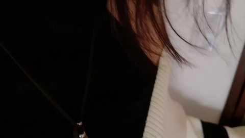 Video of a person with long, brown hair, wearing a white sweater, and a black necklace. The background is dark and indistinct, focusing on the person's upper body and neck.