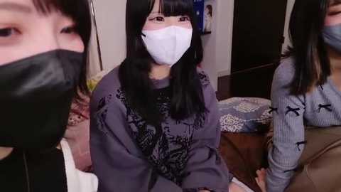 Video of three Asian women in masks and casual attire, sitting on a couch in a dimly lit living room with a TV in the background.
