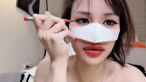Media: Video of a young Asian woman with fair skin, dark hair, and large eyes. She's applying eyeliner with a red pen, wearing a white medical mask over her mouth. Background shows a beige wall and a black and white patterned blanket.