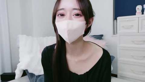 Video of an East Asian woman with long, straight black hair, wearing a white face mask, black top, and sitting in a white bedroom with a dresser and bed in the background.