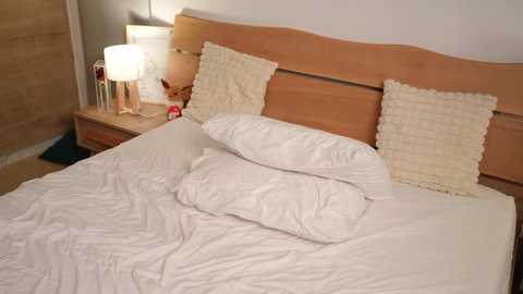 Video of a minimalist bedroom with a light wood headboard, white bed linens, two cream throw pillows, and a bedside table with a lamp, a teddy bear, and a small vase.