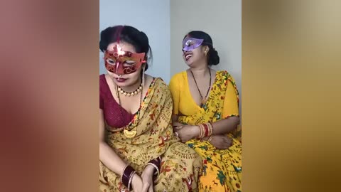 Media: Video of two Indian women in traditional sarees, one in red, the other in yellow, both wearing colorful masks and jewelry, sitting against a plain background.