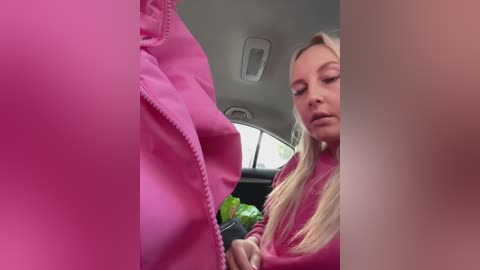Video of a blonde woman in a pink jacket, sitting in a car, with a blurred figure in the foreground.