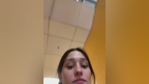 Video of a young woman with light skin and brown hair, wearing a grey top, captured from below, in a brightly lit room with beige and yellow walls and a ceiling light.