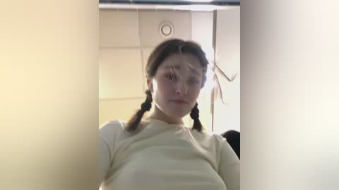 Video of a young woman with fair skin and braided hair, wearing a cream-colored top, taken from an angled, partially obscured perspective. The background shows a light-colored, tiled wall.