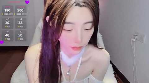 Video of a young Asian woman with long, straight brown hair, wearing a white off-shoulder dress, kneeling on a white fur rug. Background features a white wall and a purple heart overlay.