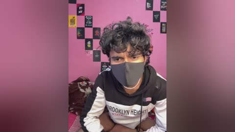 Video of a person with curly black hair, wearing a black face mask, a white and black striped hoodie, and sitting on a bed with a pink wall and various posters.