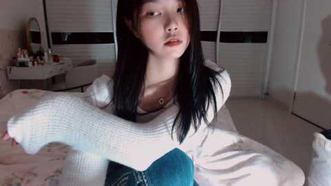 Video of a young Asian woman with long black hair, wearing a white sweater and blue jeans, hugging a large white stuffed animal in a cozy, dimly-lit bedroom with a floral bedspread and a vanity table.