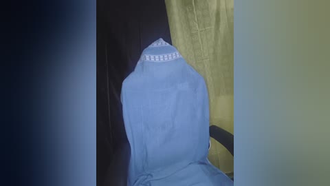 Video of a person wearing a light blue, hooded robe with white trim, standing in a dimly lit room with dark walls and a mustard-yellow curtain.
