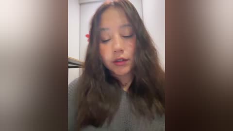 Video of a young Asian girl with long, wavy brown hair, wearing a gray sweater, with a red hair clip on the left side, eyes closed, and a serene expression.