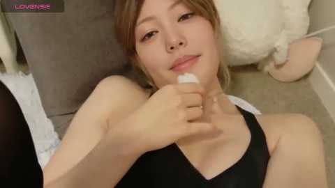 A young East Asian woman with light skin and straight, light brown hair, wearing a black tank top, sits on a gray sofa, applying lotion to her feet.