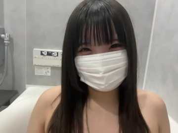 Video of an East Asian woman with long, straight black hair and a white surgical mask, wearing a white top, in a modern, sterile bathroom with a gray tiled wall and a digital shower controller.