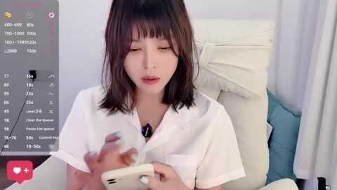 A video of a young Asian woman with straight, shoulder-length dark hair, wearing a white lab coat, sitting on a white cushion, looking at a smartphone.