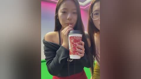 Video of an East Asian woman with long brown hair, wearing a black off-shoulder top, red skirt, and red lipstick, sipping from a red and white coffee cup, in a modern, brightly lit room with green and pink walls.