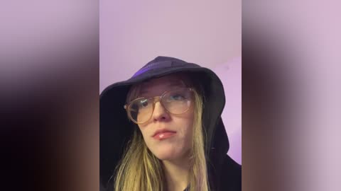 Video of a fair-skinned, blonde woman wearing a black hooded jacket and clear glasses, against a blurred, light-colored background.