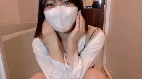 A video of an East Asian woman with straight black hair, wearing a white surgical mask and white robe, sitting in a bathtub with a white tiled wall in the background.