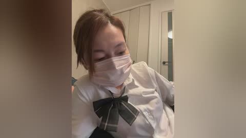 Video of an Asian woman in a white shirt with a black bow tie, wearing a face mask, in a dimly lit room with a door and light fixture in the background.