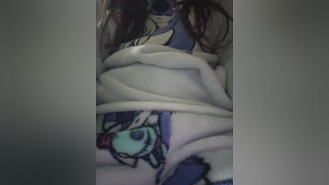 Video of a person wrapped in a white blanket featuring a cartoonish blue dog with red tongue. The person's long brown hair is visible. The background is dimly lit, suggesting a cozy indoor setting.