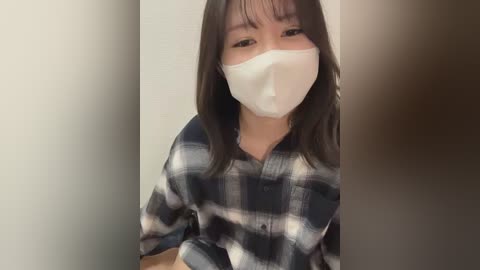 A video of an East Asian woman with long black hair, wearing a white surgical mask, blue plaid shirt, and a neutral expression, taken indoors with blurred background.