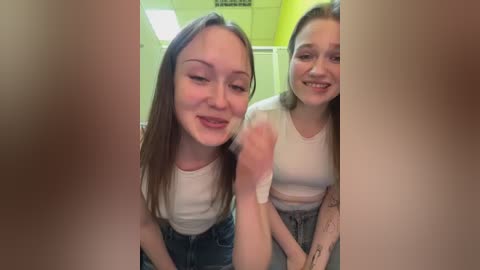 Video of two smiling young women with fair skin, wearing white crop tops and denim shorts, standing close in a brightly lit room with a yellow wall and fluorescent ceiling lights.
