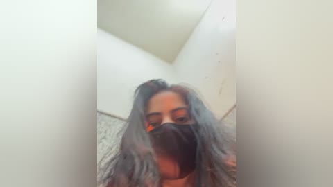 A blurred video of a young woman with long, wavy, gray hair, wearing a black face mask, standing in a small, white-tiled bathroom with a high, sloped ceiling.