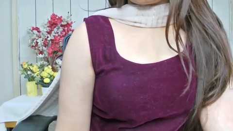 A video of a woman with long brown hair, wearing a sleeveless maroon top and beige scarf, against a background of white shutters and a floral arrangement.