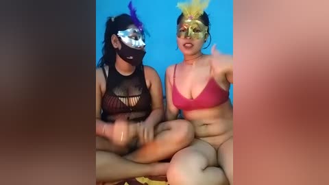 Video of two women in revealing costumes, one with a black fishnet top, the other in a red bra, both with colorful masks, sitting cross-legged against a blue background.