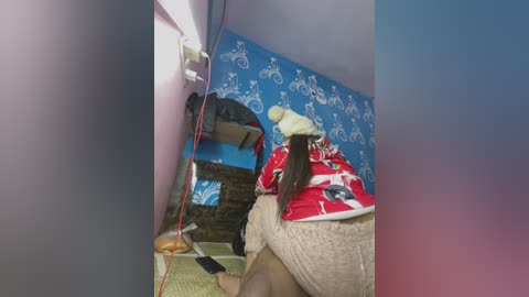 A video showing a woman in a red and white patterned dress with a beige towel, facing a mirror with a blue and white patterned background, standing in a room with a red electrical cord.