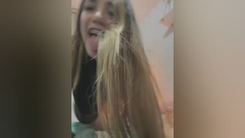 Video of a young woman with long, light brown hair sticking out of her mouth, wearing a black top, in a dimly lit room.