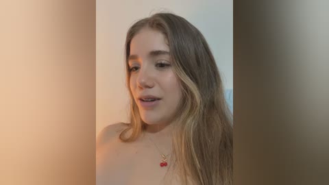 A video of a young woman with fair skin and long, light brown hair, wearing a red necklace, smiling softly. The background is a blurred gradient of beige and light green.