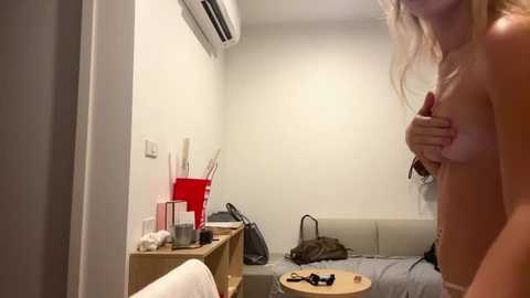 Video of a topless blonde woman, covering her breasts with her hands, standing in a sparsely furnished, dimly lit hotel room with a bed, red chair, and air conditioning unit on the wall.