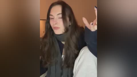 Video of a smiling woman with long, wavy dark hair, wearing a dark scarf and navy sweater, making a peace sign, indoors with warm lighting.
