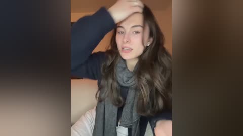 A video of a young woman with long brown hair, wearing a dark blue sweater and grey scarf, wiping her forehead with her right hand. Background shows a blurred indoor setting.