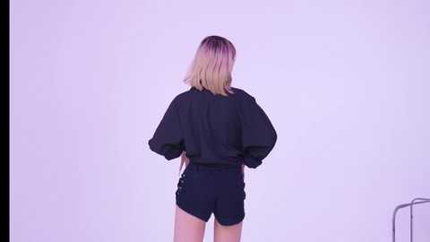 Video of a woman with shoulder-length blonde hair wearing a black, oversized, long-sleeved shirt and black shorts against a plain white background.