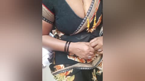 A video of a woman with light brown skin, wearing a black saree with floral patterns, a deep V-neck blouse, and a gold necklace. She holds a black purse and has multiple bracelets on her wrist.