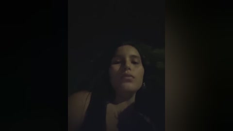 Video of a woman with fair skin and dark hair, eyes closed, wearing a brown top, in a dimly lit room with dark walls, creating a moody, intimate atmosphere.